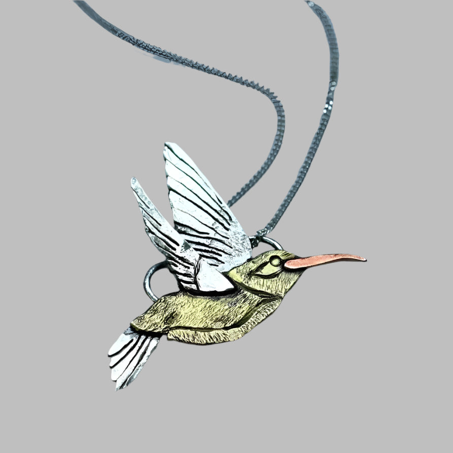 Hand fabricated silver, gold and copper hummingbird necklace with chain on a grey background.