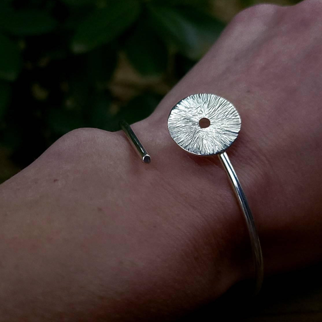 Silver Cuff Manifestation bracelet with circle with lines on a woman's wrist in front of green leaves.