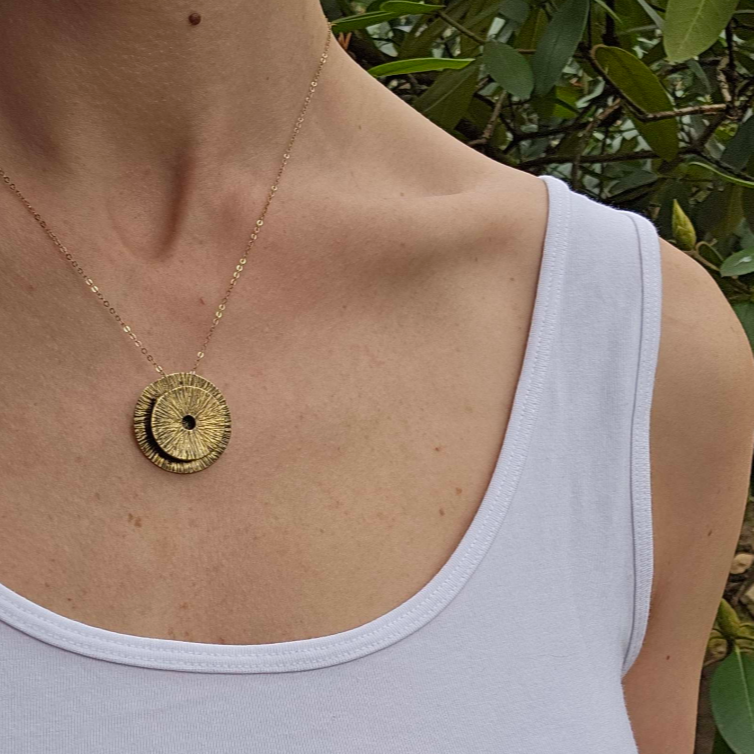 Woman wearing a 3 dimensional gold circle necklace with white sleeveless top in front of branches with green leaves