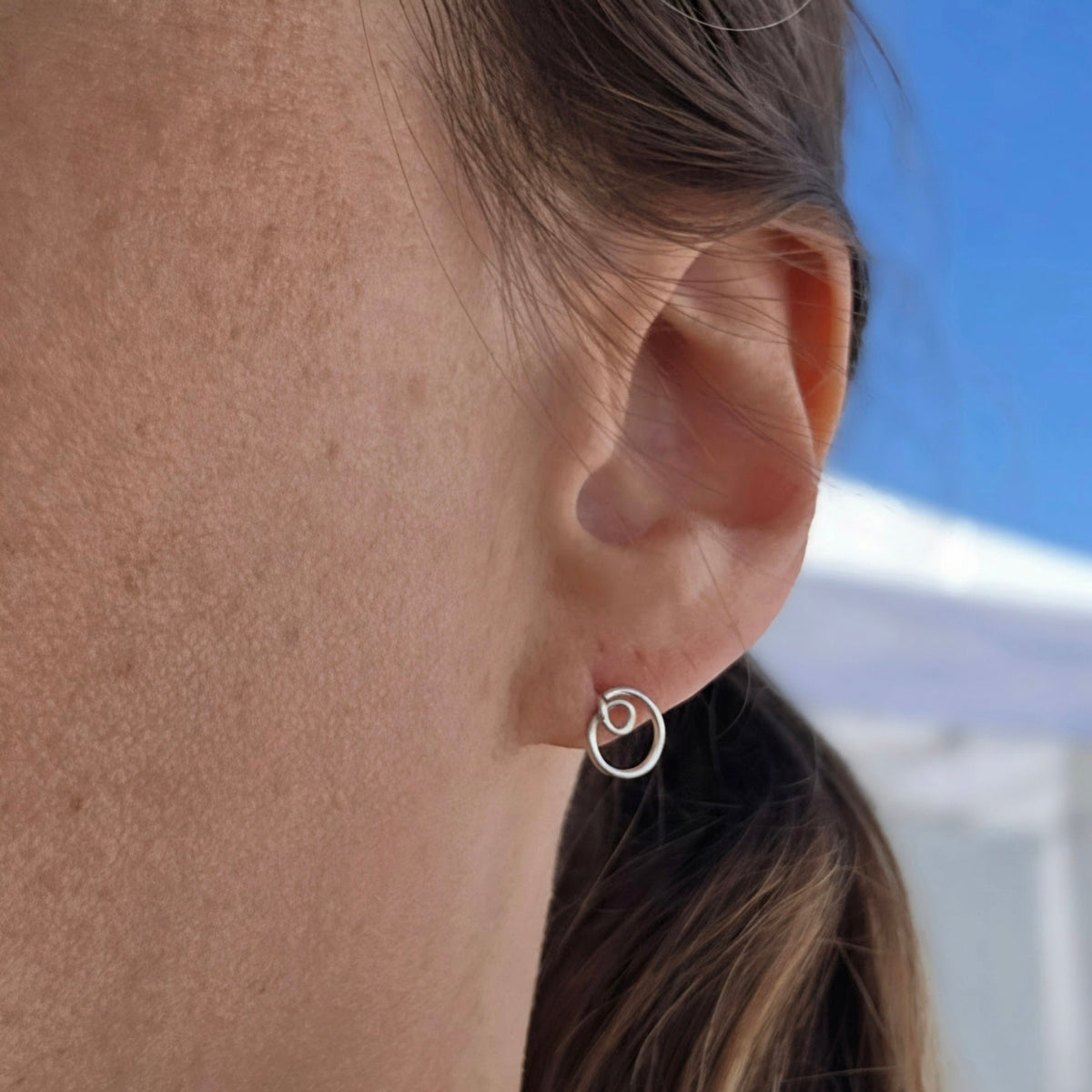 A close-up of a person's ear wearing a small silver spiral hoop earring, with hair and skin visible against an outdoor blue sky and white tent background.