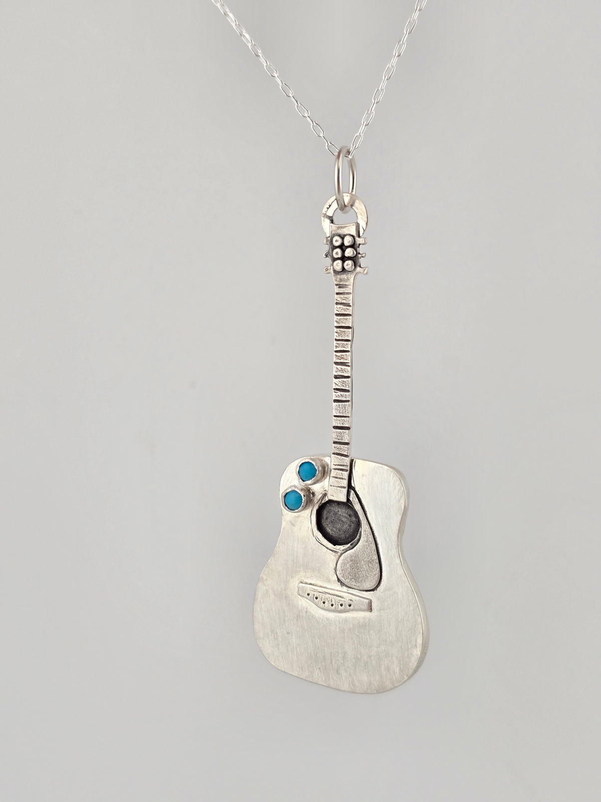 Handmade silver guitar necklace hanging in front of a grey background. 