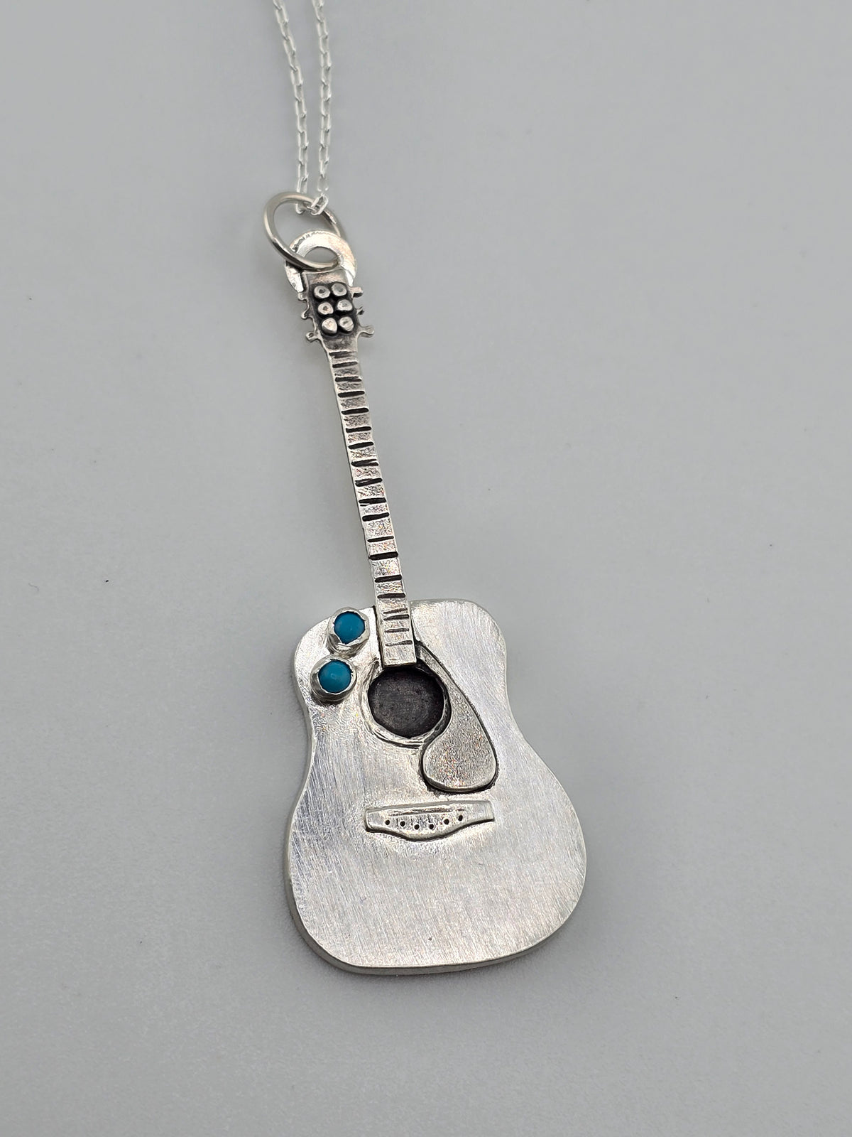 Close up shot of the details of a fully handmade silver guitar necklace with turquoise accent stones made by Jaclyn Nicole. 
