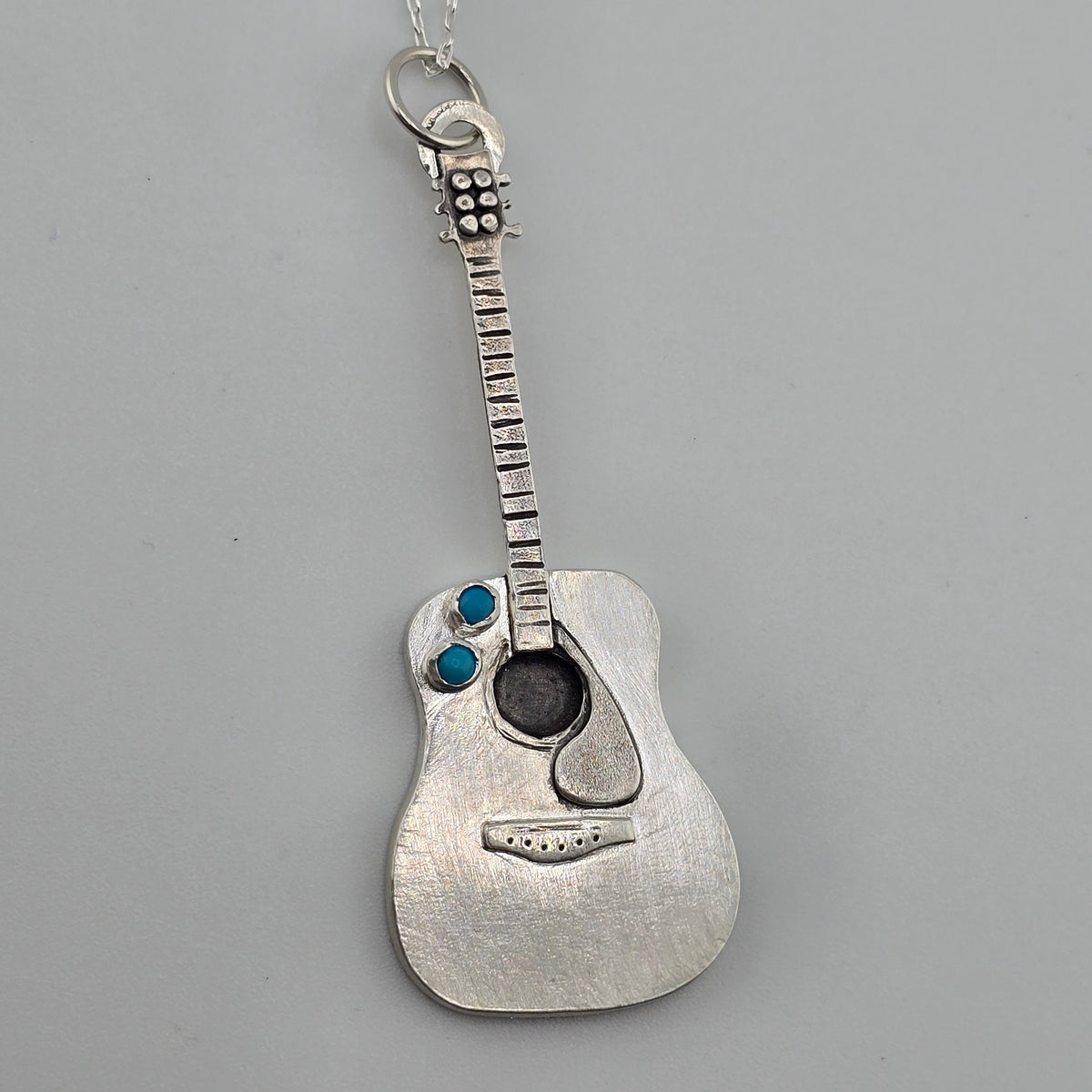 Handmade silver guitar necklace with turquoise stone accents on a grey background. 