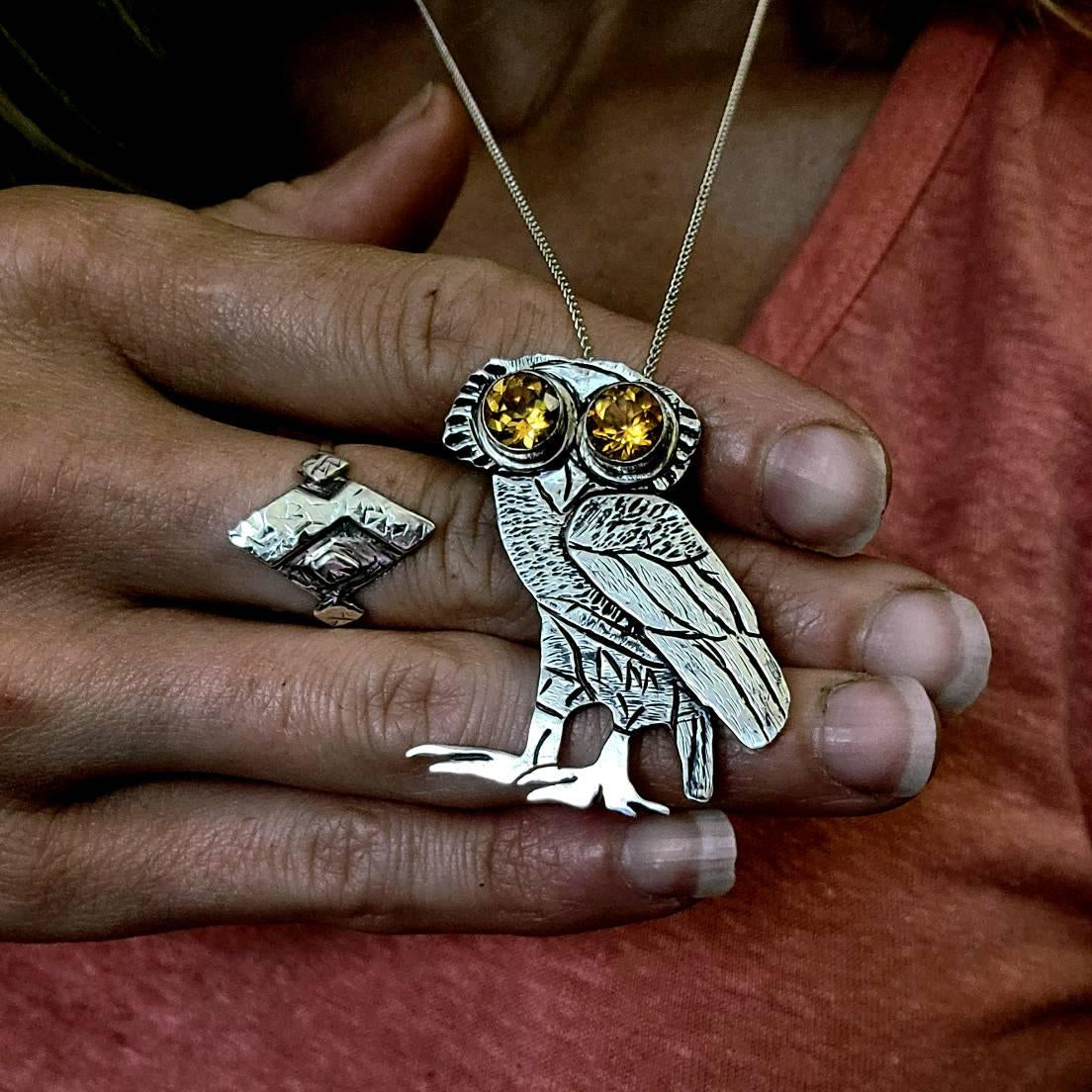Woman in orange shirt with hand in front of her wearing ring and holding forward handmade athena owl necklace.