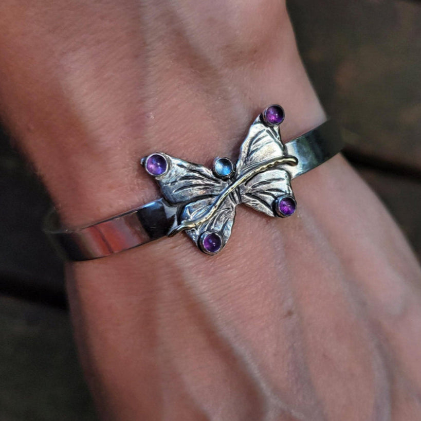 close up of handmade one of a kind butterfly on a leaf pendant necklace set with aquamarine and amethyst stones displayed on woman's wrist in front of wood slats