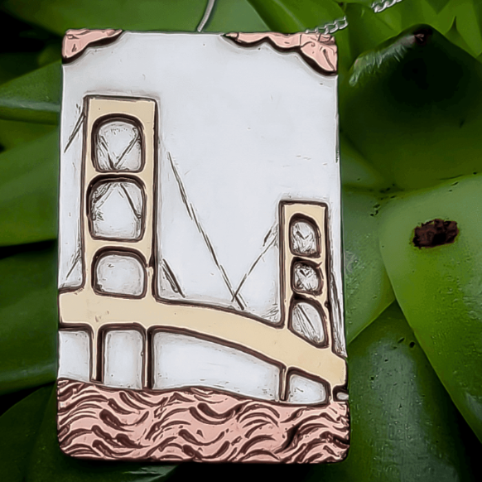 Handmade custom jewelry necklace of Mackinac bridge in Michigan in silver, gold and copper atop green leaves